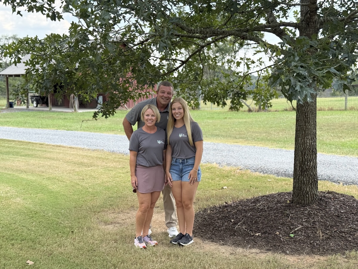 Three people wearing matching gray shirts stand smiling under a large tree on a rural property.