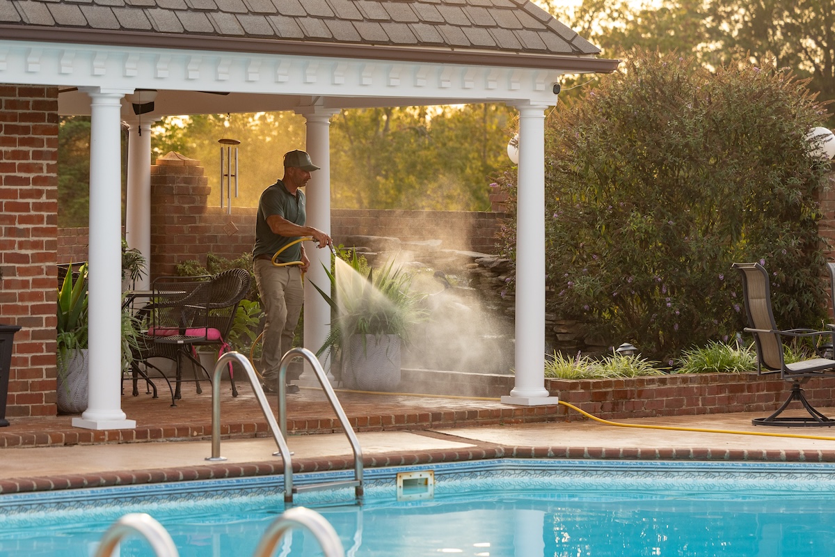 A man mists plants on a sunny patio by a swimming pool, with golden