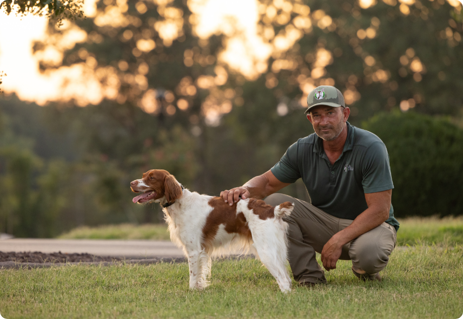 A man in a green shirt and cap crouches, petting his brown and white Brittany span