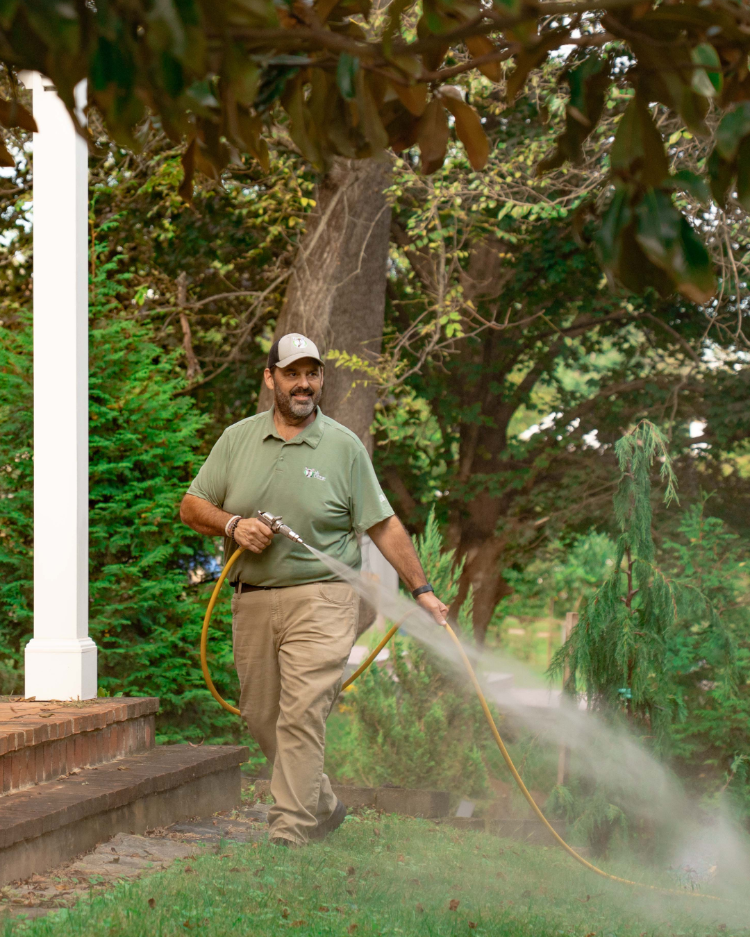 Smiling technician in uniform sprays a residential lawn with a pest control hose.