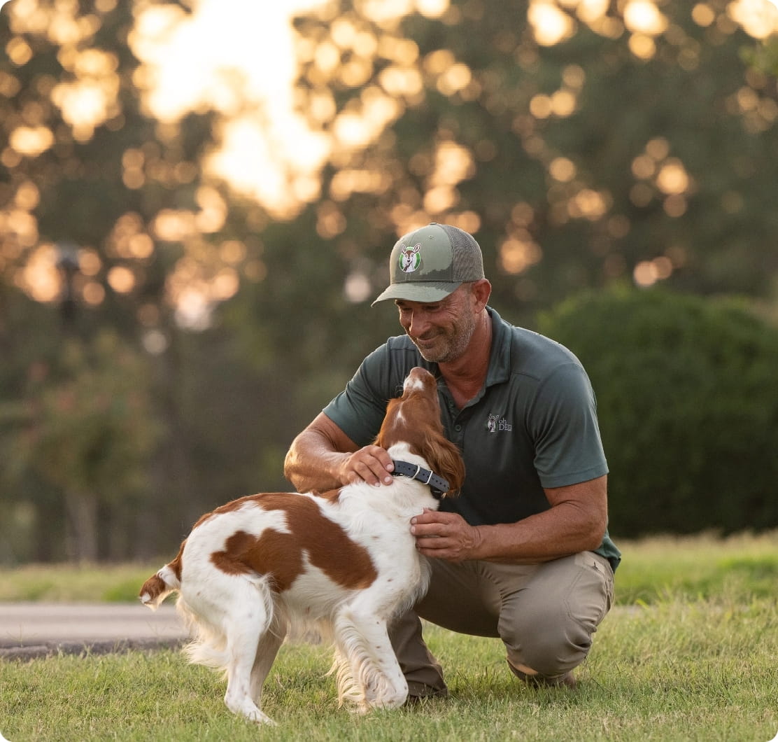 A smiling man in a green cap lovingly pets his brown and white dog on a sunny