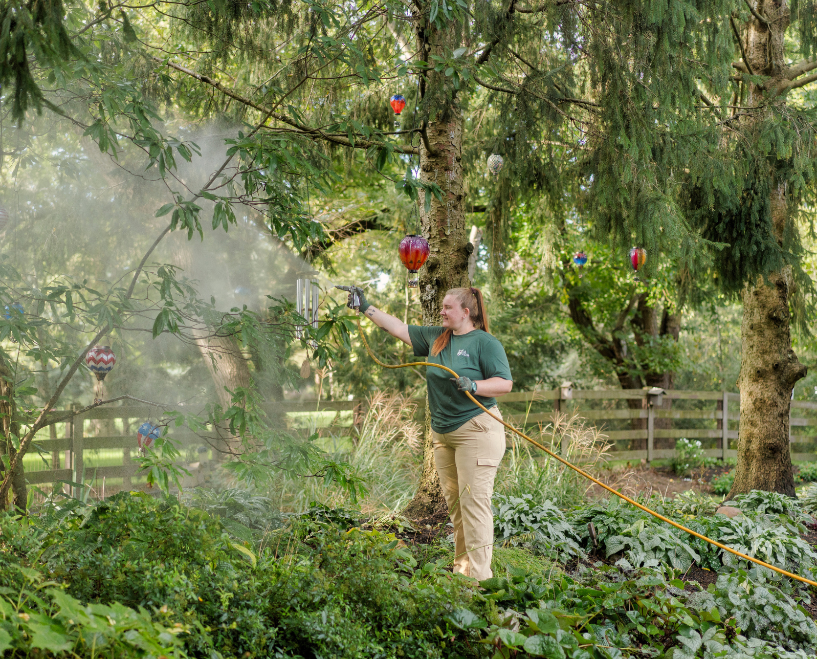 A woman mists garden plants and decorative hot air balloons hanging from trees.