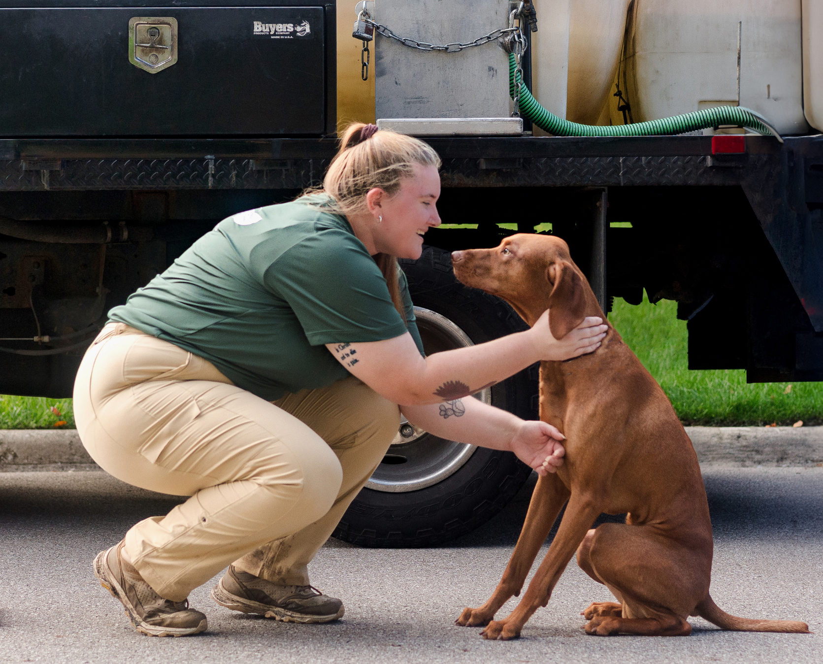 A smiling woman in a green shirt crouches to lovingly pet a reddish-brown Vizsla dog