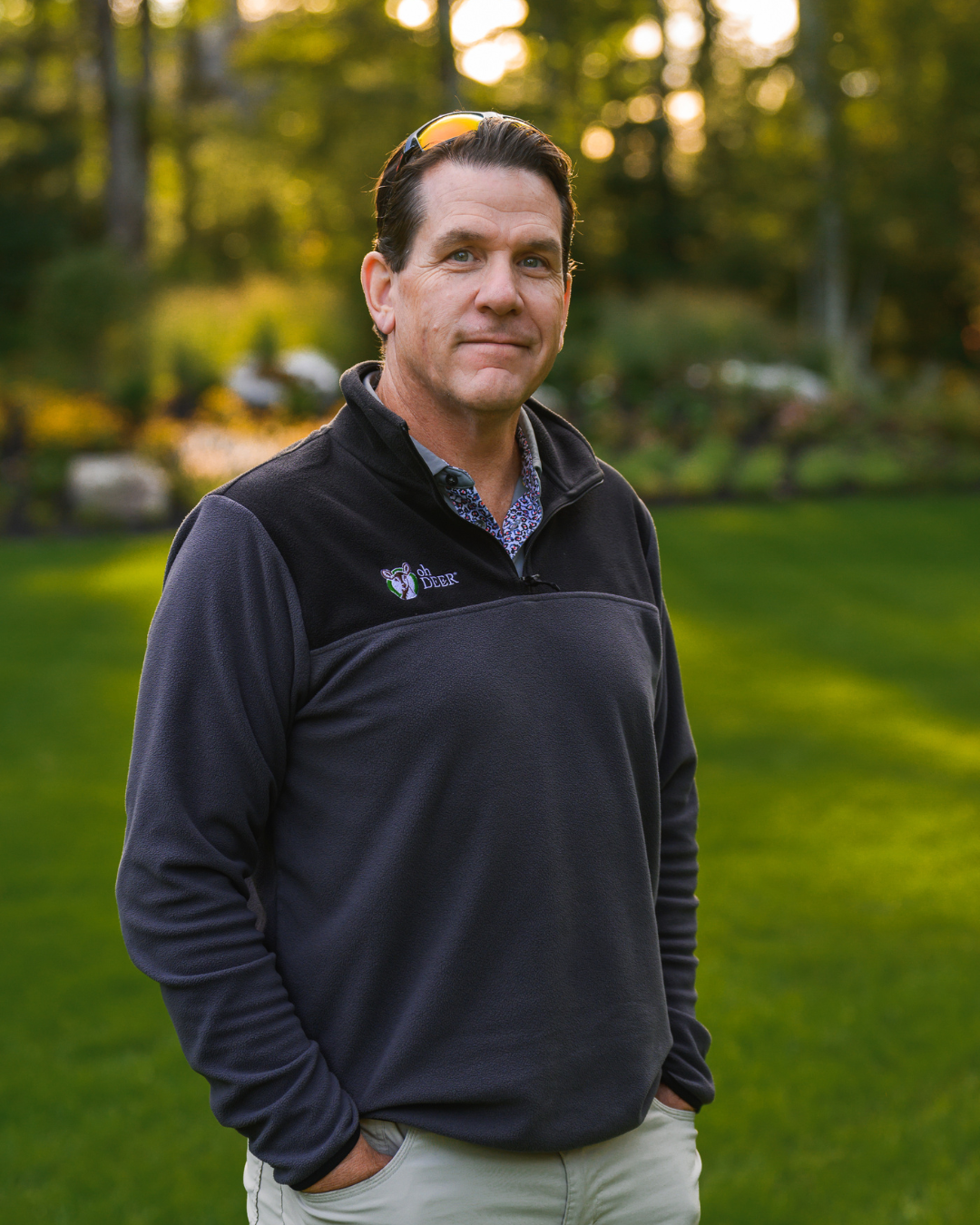 A smiling man in a fleece pullover and sunglasses stands in a sunny, green outdoor setting.