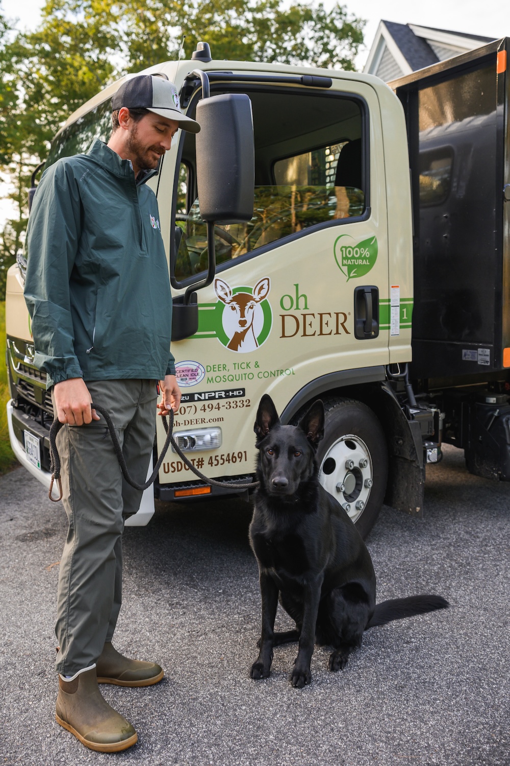 A man in a green jacket and cap stands with a black dog beside an 'Oh Deer
