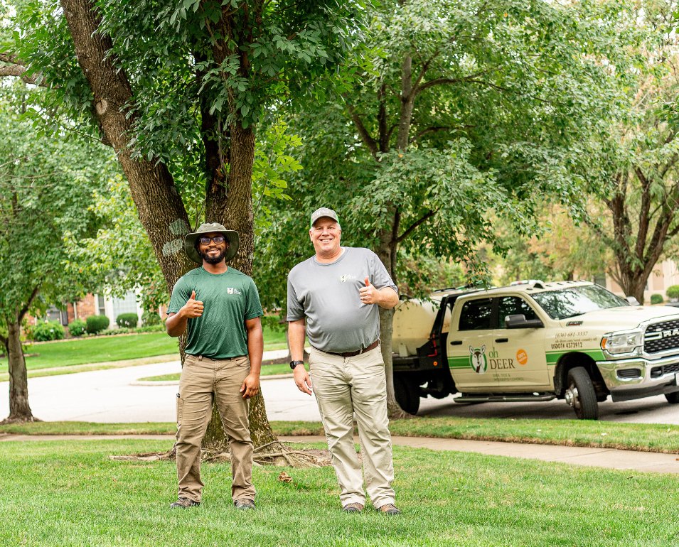 Smiling Oh Deer technicians give thumbs-up on a lawn with their branded service truck nearby.