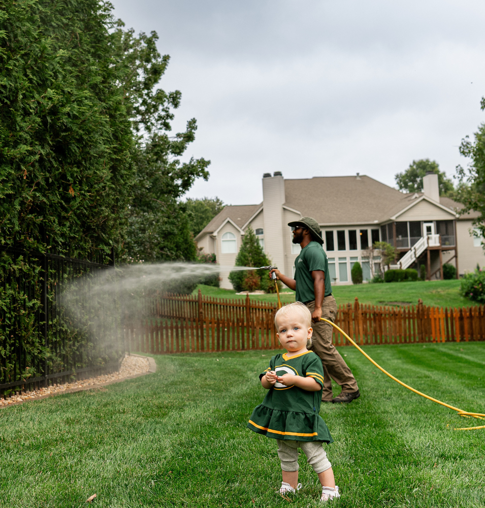 A young child in a green Packers dress stands in a lush backyard while a landscaper