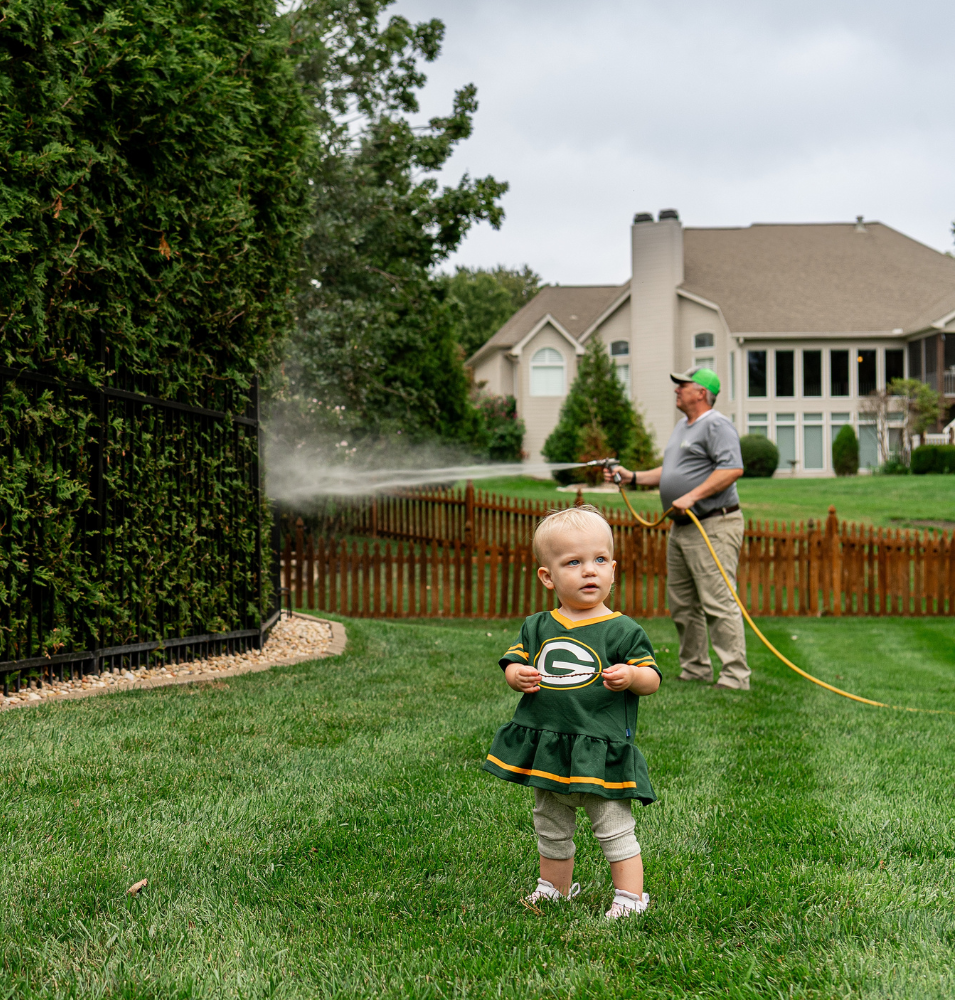 A toddler in a Green Bay Packers outfit stands on a lush lawn as a man waters a