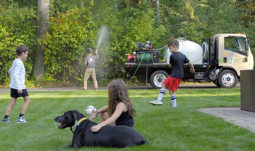 ohDEER owner spraying all-natural tick & mosquito control in yard with kids playing soccer and dog