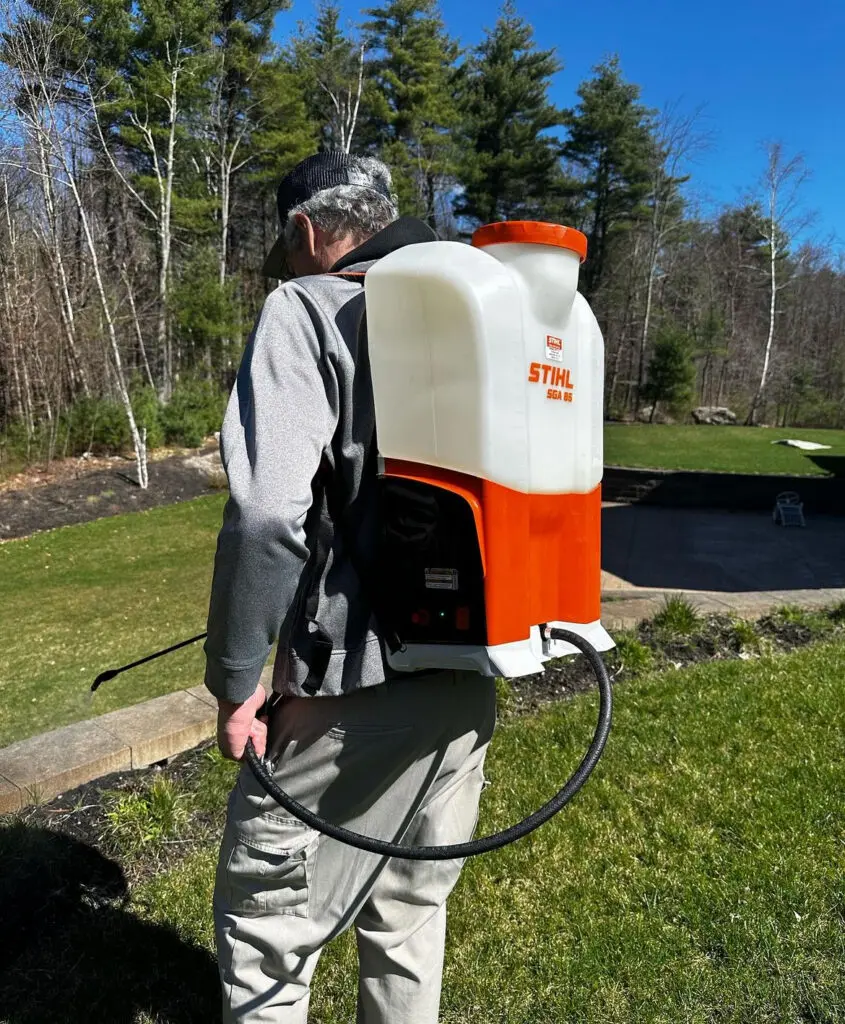 A person wears a Stihl SGA 85 backpack sprayer on a sunny lawn with trees