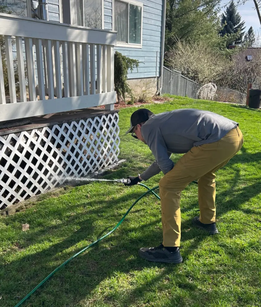 A person pressure washes the white lattice skirting beneath a blue house's deck.