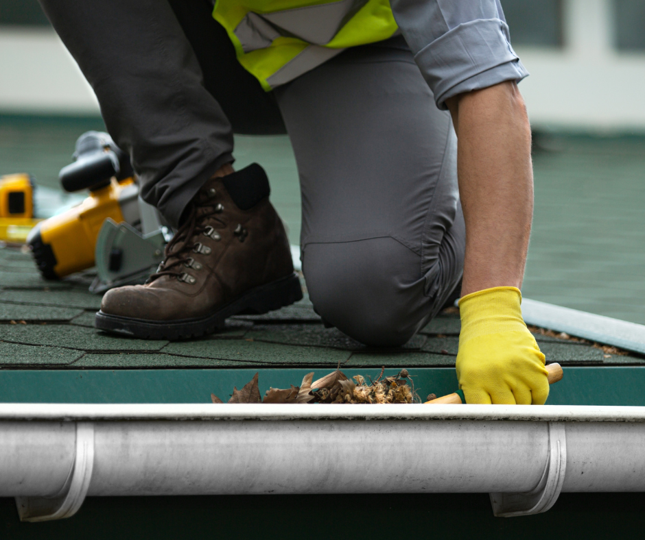 A worker in safety gear cleans leaves and debris from a roof gutter.