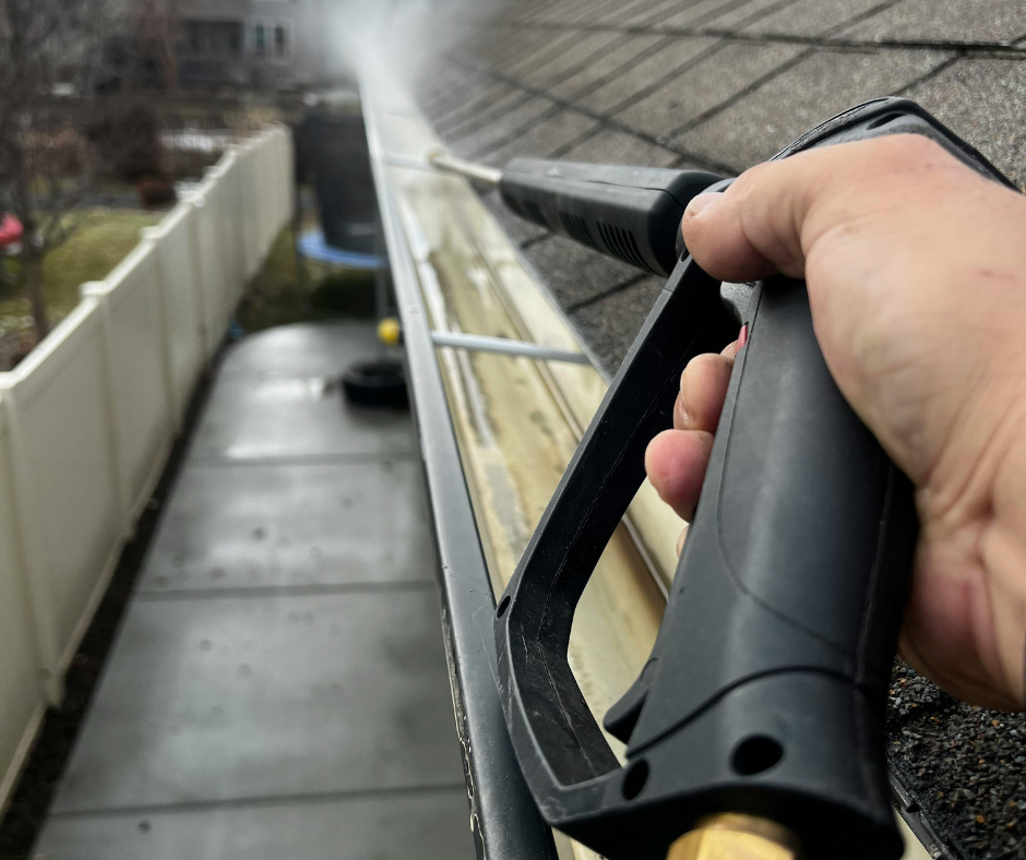 A person uses a pressure washer to clean a dirty roof gutter, spraying water to