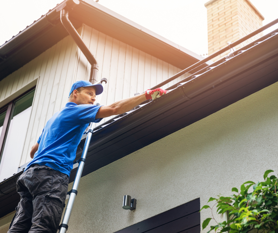 A man in a blue uniform cleans dark gutters on a house while on a ladder. TITLE