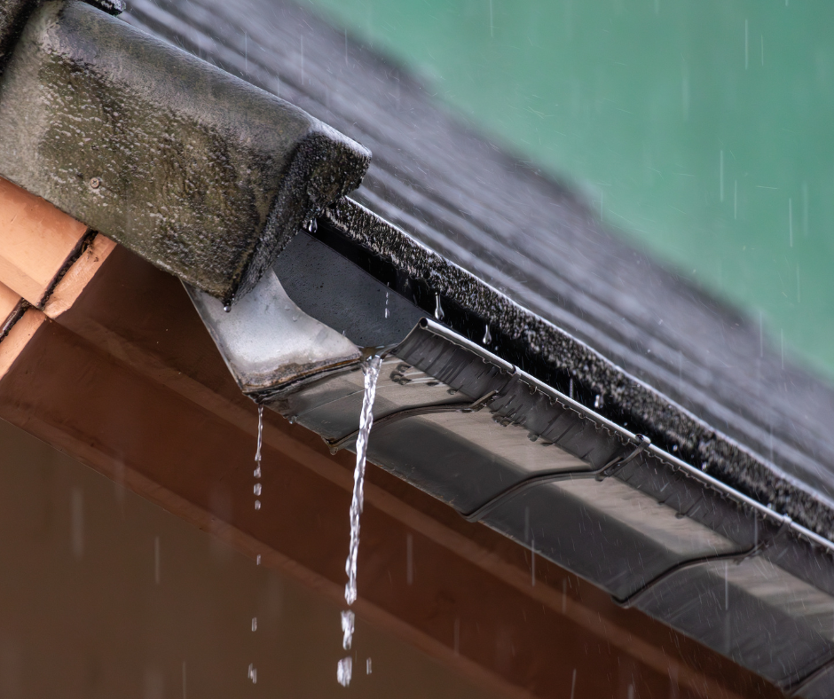 Rainwater streams from a dark roof into an overflowing metal gutter system with terracotta tiles