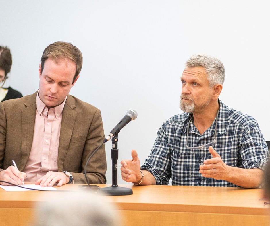 A man with a gray beard speaks into a microphone while another man writes notes at a table.