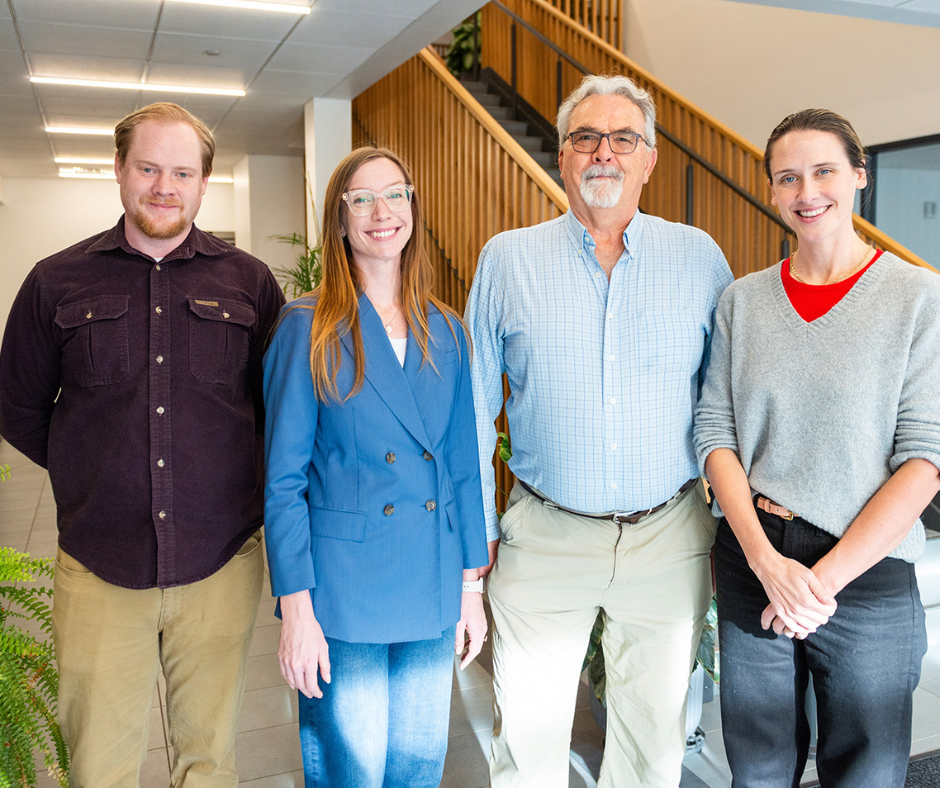 Four smiling professionals, two men and two women, stand together in a modern office.