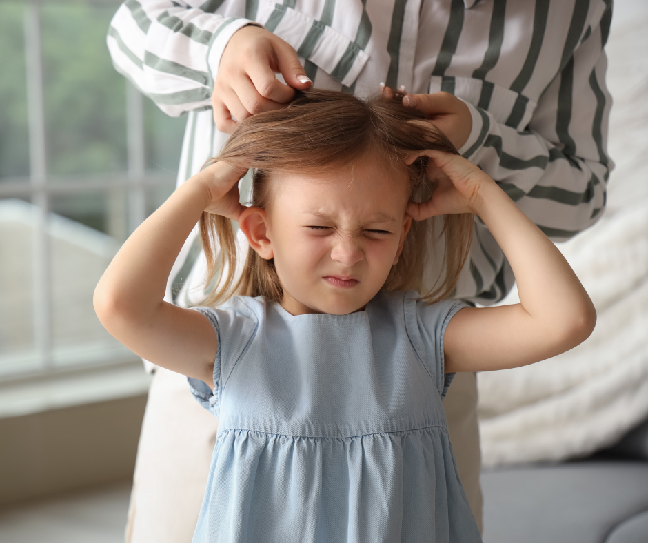 A young girl with an uncomfortable expression scratches her head as an adult checks her hair for ticks 