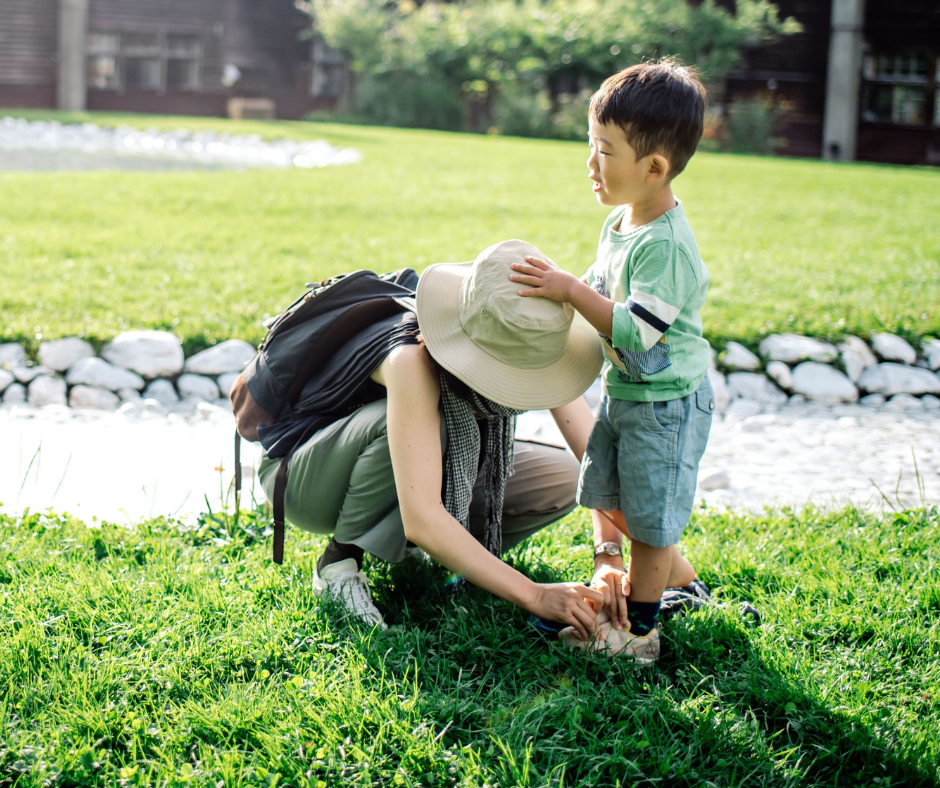 A woman crouches to help a young boy with his shoe on a sunny grassy lawn as