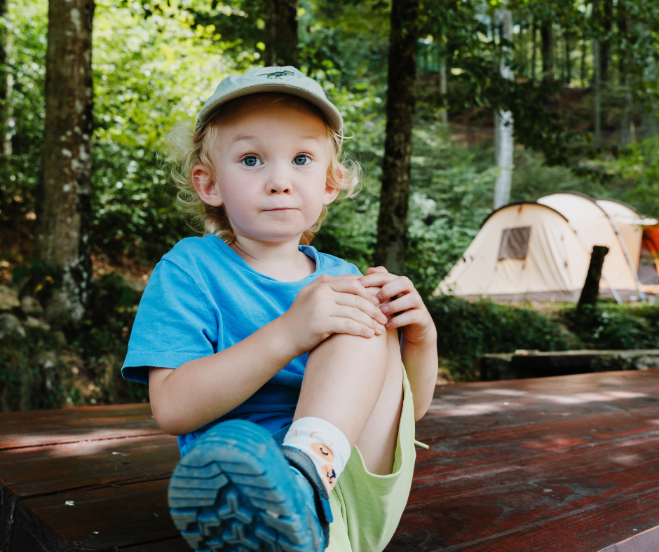 A curious blonde child in a baseball cap sits on a picnic table at a forest campsite with