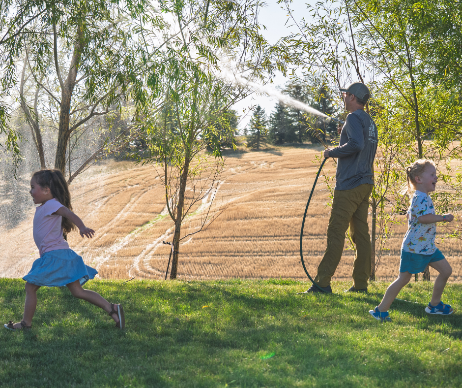 A man waters plants while two happy children run through the refreshing spray on a sunny day