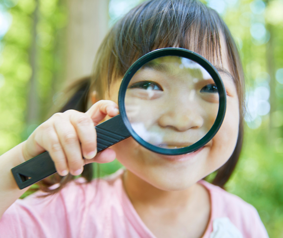 A smiling young child holds a magnifying glass to her face, magnifying her eyes and nose.
