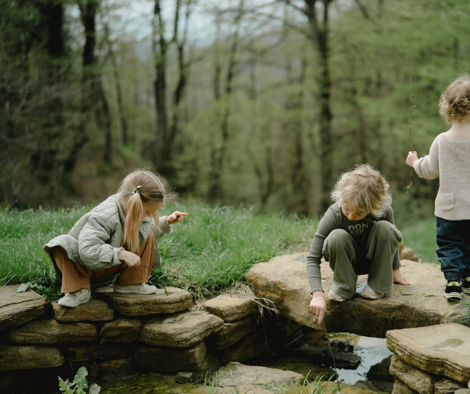 Three children play with sticks in a small stream surrounded by green foliage and stone steps